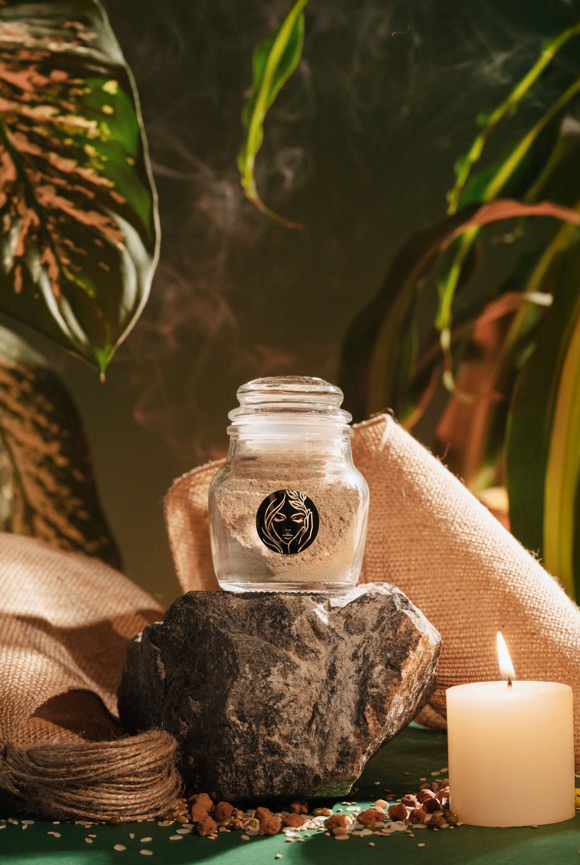 Glass jar of Prakriti's Face Pack with a lid on a rock, featuring a Prakriti Logo, with a blurred natural background.